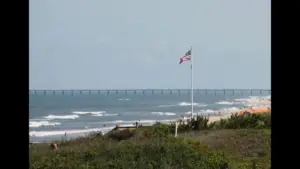 A view of a beach with waves, a flagpole displaying the American flag, and a distant pier. Corolla, NC, USA, Labor Day weekend destinations