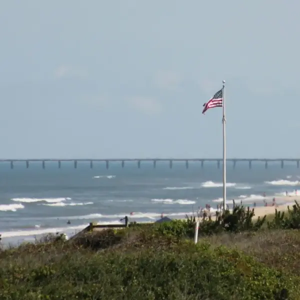 A view of a beach with waves, a flagpole displaying the American flag, and a distant pier. Corolla, NC, USA, Labor Day weekend destinations