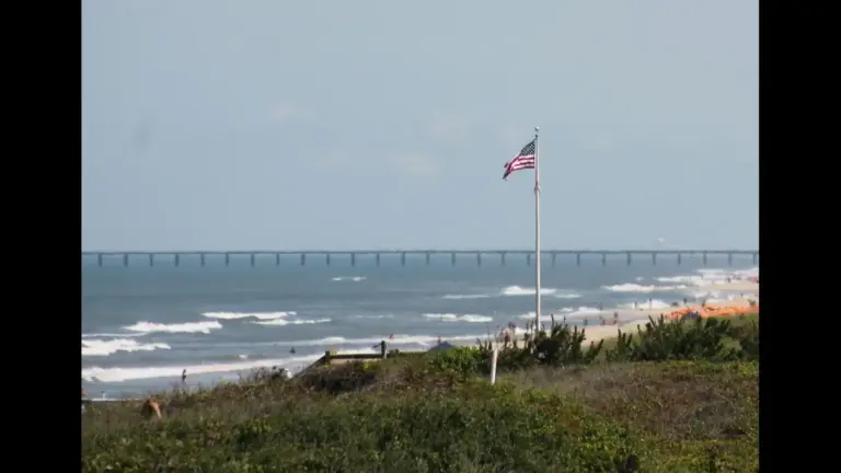 A view of a beach with waves, a flagpole displaying the American flag, and a distant pier. Corolla, NC, USA, Labor Day weekend destinations