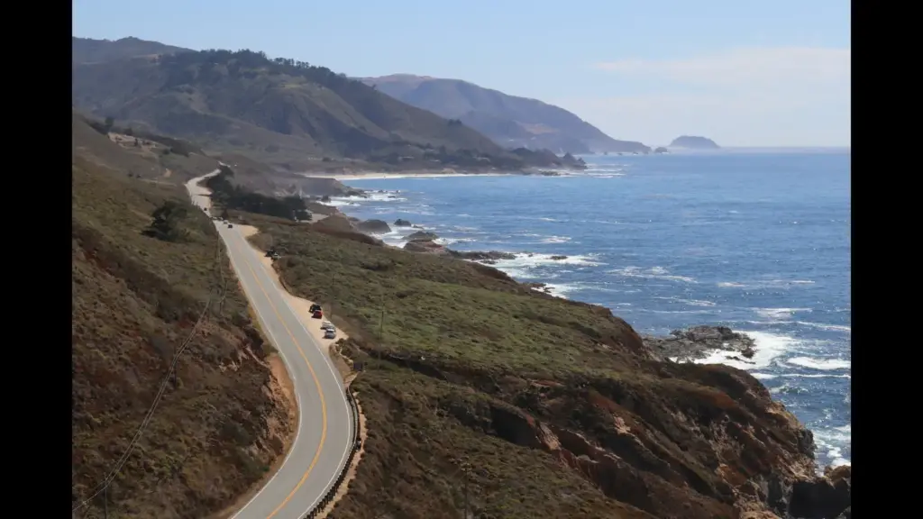A scenic coastal view with a winding road alongside the ocean, featuring rolling hills and distant mountains. Big Sur, CA, Pacific Coast Highway, road trip