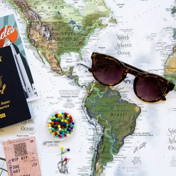 A flat lay of a world map featuring sunglasses, a U.S. passport, colorful push pins, some papers, and a ticket. Labor Day, vacation planning, trip planning, summer vacation