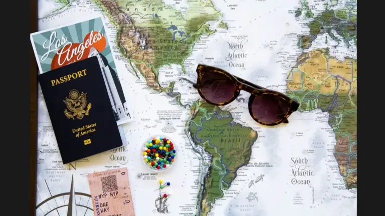 A flat lay of a world map featuring sunglasses, a U.S. passport, colorful push pins, some papers, and a ticket. Labor Day, vacation planning, trip planning, summer vacation