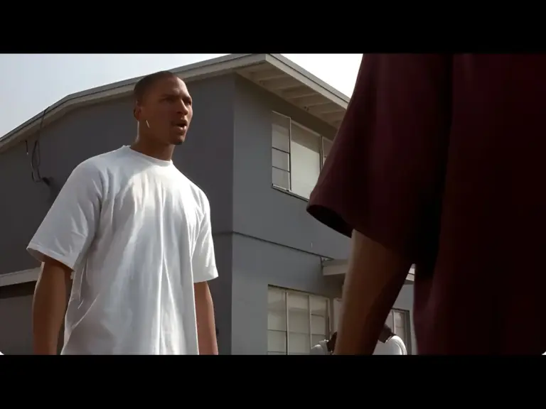 A scene from "Menace II Society": a dramatic confrontation between two young men in an urban setting. One is in a white T-shirt and appears to be speaking assertively, while the other, wearing a dark shirt, stands facing him. They are surrounded by a group of people, and a residential building is visible in the background.