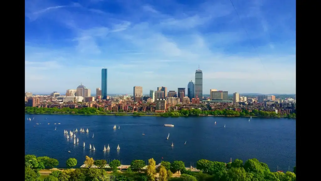 A panoramic view of a city skyline alongside a river, featuring modern skyscrapers and sailboats on the water, with a clear blue sky. Boston, MA, USA