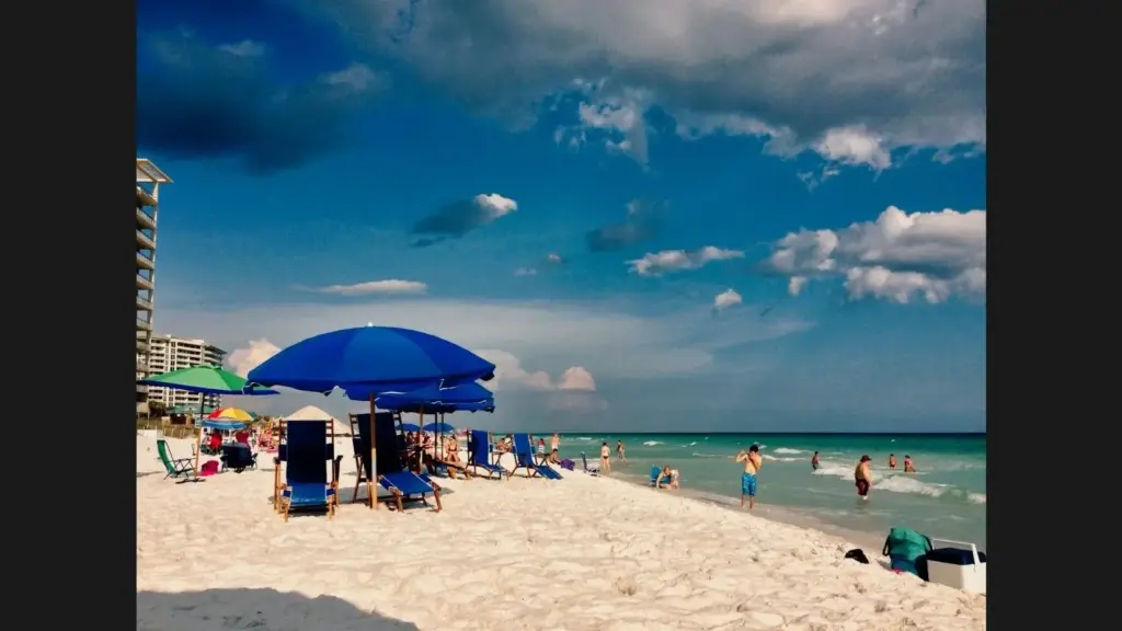 A beach scene with umbrellas, lounge chairs, and people enjoying the water under a cloudy sky. Destin, FL, USA