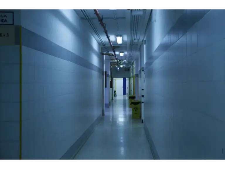 Dimly lit hospital corridor with bluish fluorescent lighting, yellow bins along the wall, and a faint haunted call light glowing near the end of the hallway.