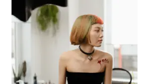Stylish woman with colorful bob haircut and shoulder tattoo sits in a bright room, embodying the soft girl aesthetic with modern flair.