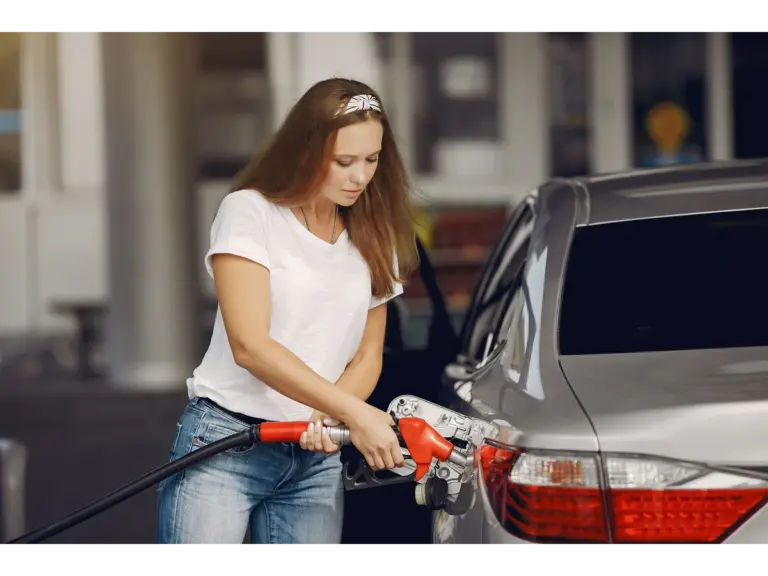 Woman in casual attire refueling a car at a gas station on a sunny day, reflecting everyday travel impacted by changing gas prices.