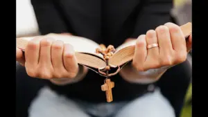 In daily devotional, someone clings to the Bible as a cross dangles from its center., showing adherence to the Word.