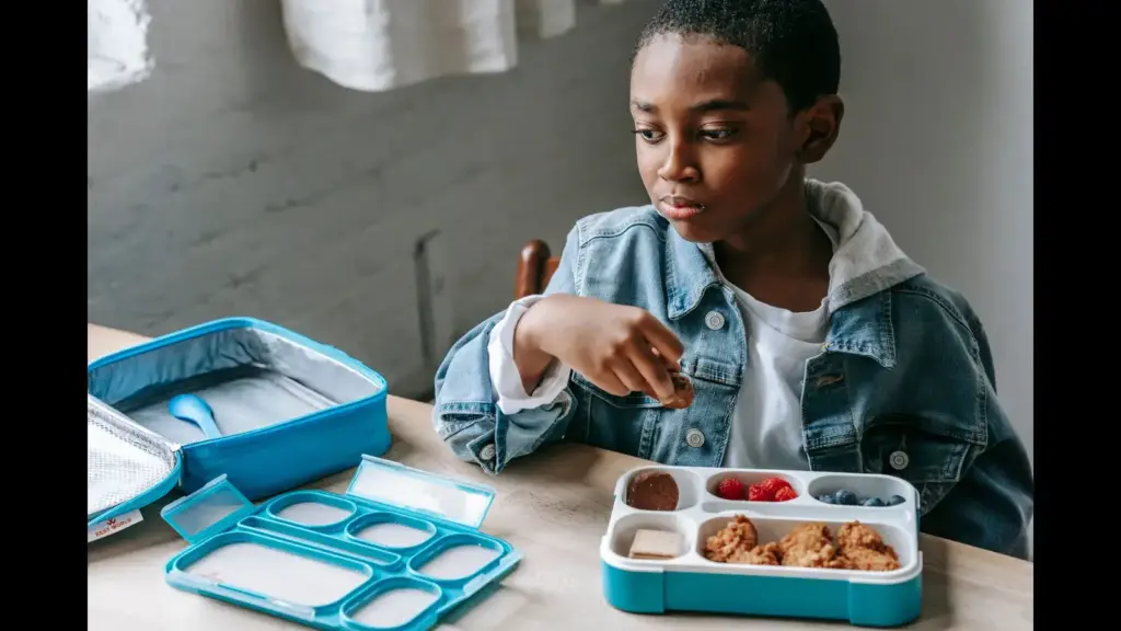 A young boy sitting at a table with a lunchbox, looking thoughtfully at his food. insulated lunch box, school lunch, back-to-school