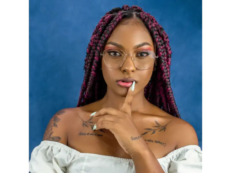 Confident woman with pink and purple braids, glasses, and shoulder tattoos poses against a blue background, embodying the soft girl aesthetic.