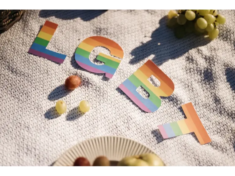 Rainbow‑colored letters spelling “LGBT” rest on a textured white blanket beside fruit, symbolizing pride and inclusion connected to Jay Lee’s story.
