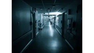 Dimly lit hospital corridor with an Exit sign, featuring a wheelchair in the distance and a haunted call light glowing faintly above a doorway.