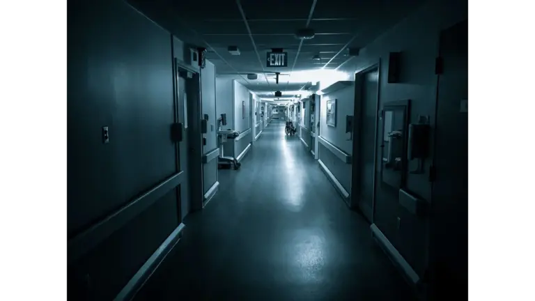 Dimly lit hospital corridor with an Exit sign, featuring a wheelchair in the distance and a haunted call light glowing faintly above a doorway.
