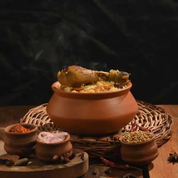 A steaming clay pot filled with rice and chicken sits on a wooden table surrounded by spices and small earthen bowls, capturing the rustic essence of ancient food and traditional cooking.