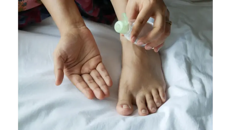 A person applies a clear liquid from a small bottle onto their bare foot resting on a white surface, showcasing a soothing foot serum self‑care moment.
