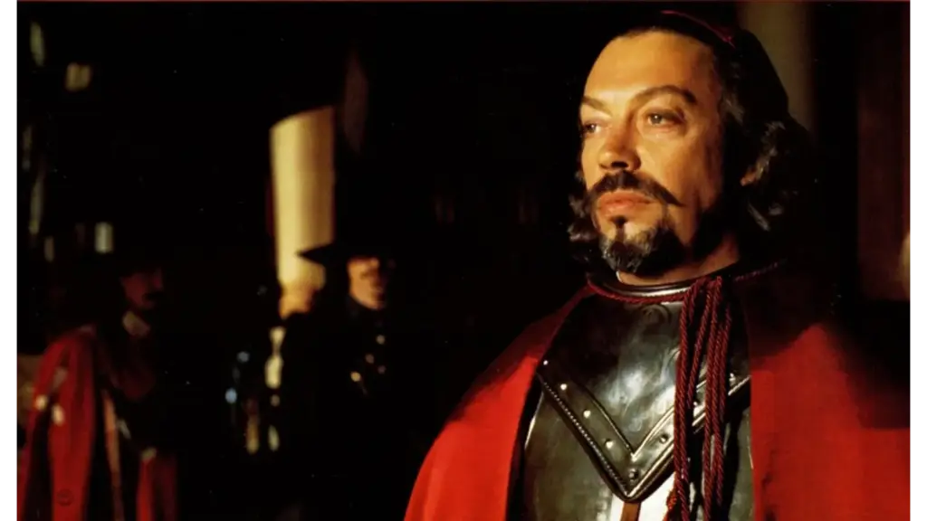 Tim Curry as Cardinal Richelieu standing stoically with Michael Wincott as Rochefort in the distance. Tim Curry wears black armor and a red cloak stands in a dim hall, his mustache and beard lit dramatically, creating a tense mood.