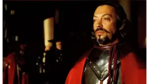 Tim Curry as Cardinal Richelieu standing stoically with Michael Wincott as Rochefort in the distance. Tim Curry wears black armor and a red cloak stands in a dim hall, his mustache and beard lit dramatically, creating a tense mood.