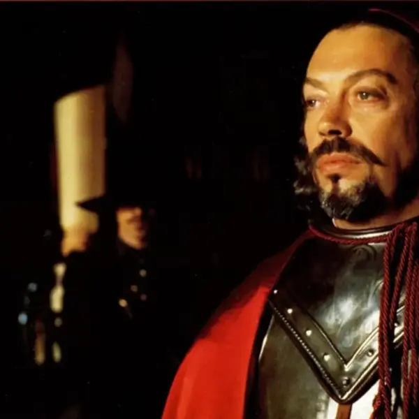 Tim Curry as Cardinal Richelieu standing stoically with Michael Wincott as Rochefort in the distance. Tim Curry wears black armor and a red cloak stands in a dim hall, his mustache and beard lit dramatically, creating a tense mood.