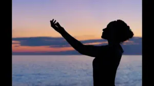 A silhouette of a person with curly hair, reaching out towards a sunset over the ocean. summer fasting and prayer traditions, late summer religious practices