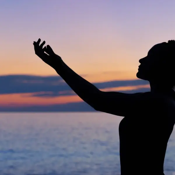 A silhouette of a person with curly hair, reaching out towards a sunset over the ocean. summer fasting and prayer traditions, late summer religious practices