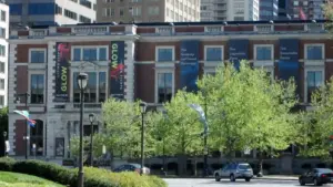 Academy of Natural Sciences of Drexel University -sunlit city street lined with leafy trees and parked cars, facing a brick building with colorful banners; a calm, bustling downtown scene.