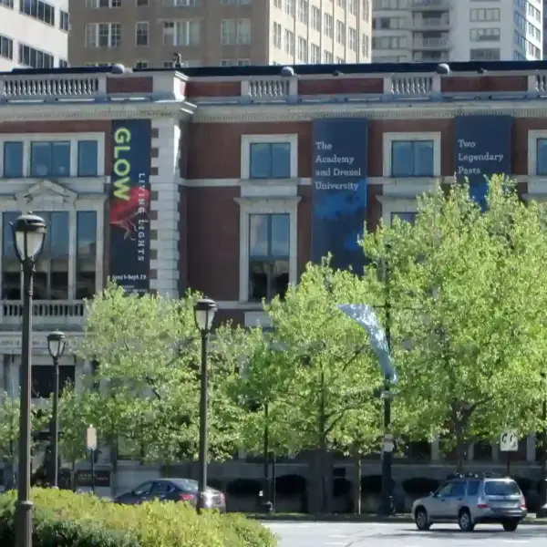 Academy of Natural Sciences of Drexel University -sunlit city street lined with leafy trees and parked cars, facing a brick building with colorful banners; a calm, bustling downtown scene.
