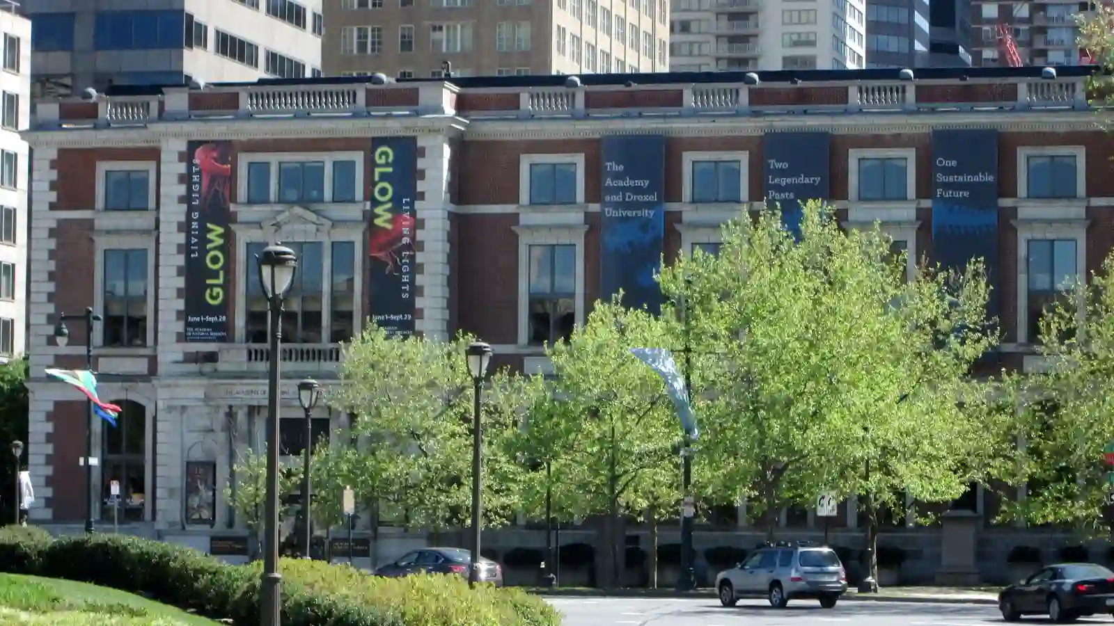 Academy of Natural Sciences of Drexel University -sunlit city street lined with leafy trees and parked cars, facing a brick building with colorful banners; a calm, bustling downtown scene.