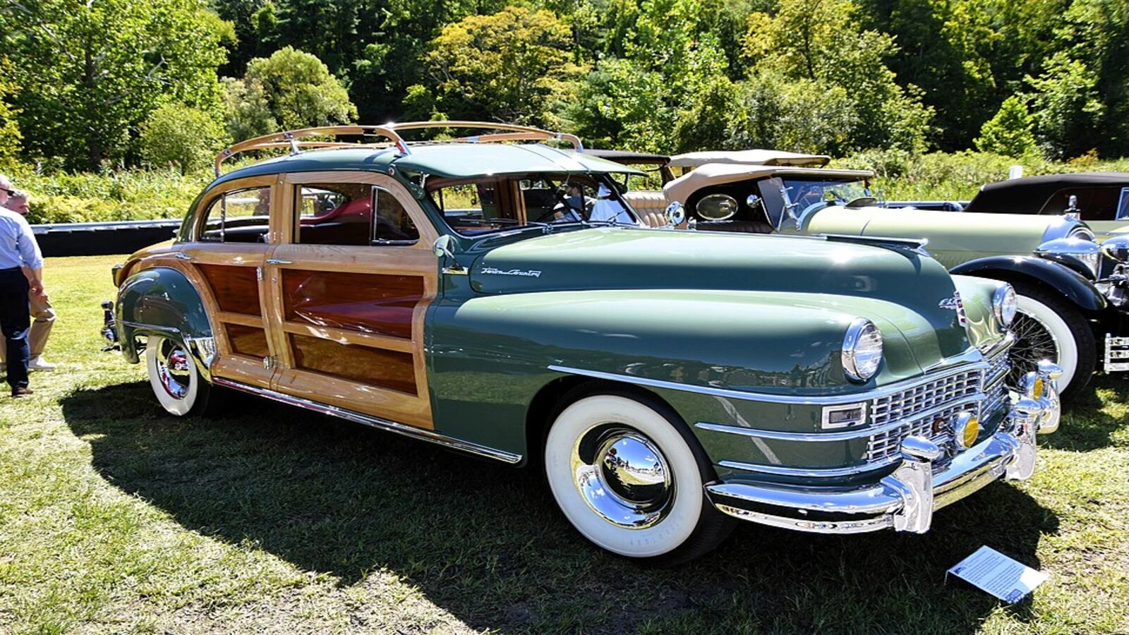 1947 Chrysler Town and Country: A vintage green car with wooden paneling parked on grass, surrounded by trees.