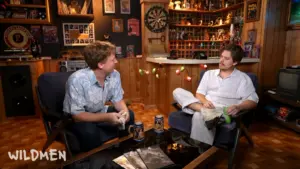 Two young men sitting in a cozy, retro-themed room with wooden paneling, surrounded by various decorations, including a dartboard and shelves filled with collectibles. One man is holding something in his hands, while the other is looking at him. They both have casual attire and are engaged in conversation. Wildmen, Dylan Sprouse, Brendan Columbus