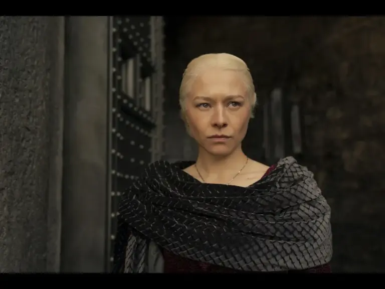 Emma D'Arcy in House of the Dragon (2022), with light hair and a serious expression, stands outside a stone structure, wearing a dark, textured shawl.