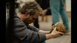 A person crouches and examines a skull with a focused expression, surrounded by blurred figures in the background. Andrew Scott in Elsinore (2026) Courtesy of Focus Features