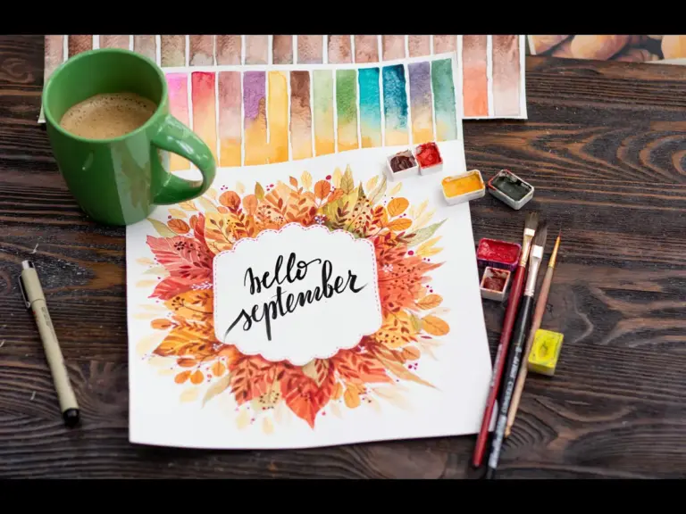 A workspace with a cup of coffee, watercolor paints, brushes, and a floral artwork saying “Hello September” surrounded by autumn leaves.