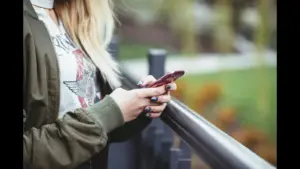 A person wearing a green jacket is holding a pink phone while standing near a railing. text messaging, relationships, texting boundaries while dating