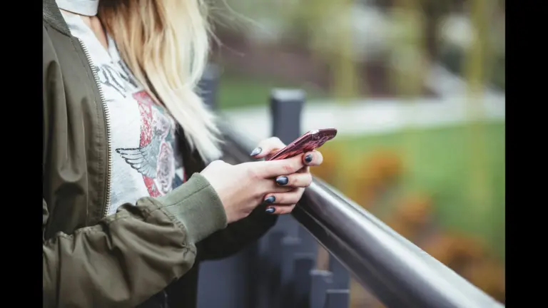 A person wearing a green jacket is holding a pink phone while standing near a railing. text messaging, relationships, texting boundaries while dating