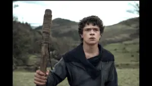 A young man poses in a field, holding a wooden stick, with greenery and trees visible in the distance. Noah Jupe in The Death of Robin Hood - Noah Jupe casted in Avengers: Secret Wars