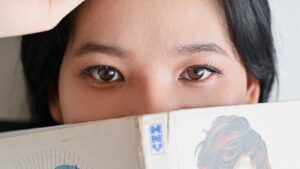 A young woman peeks over the top of an open manga, the kind of shojo romance She Loves Lies Too Much fans are waiting on