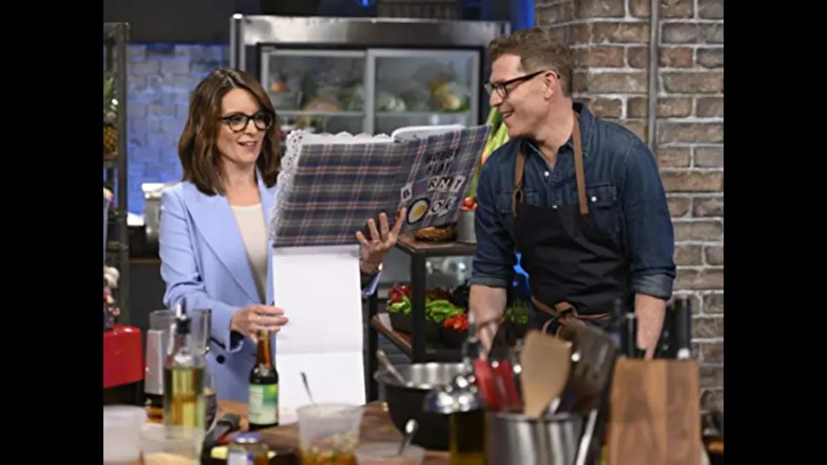 A woman in a blue blazer reading a large recipe book while a man in an apron looks on, both in a kitchen setting with various cooking ingredients and equipment around them. Tina Fey and Bobby Flay in Fey vs. Flay (2019)