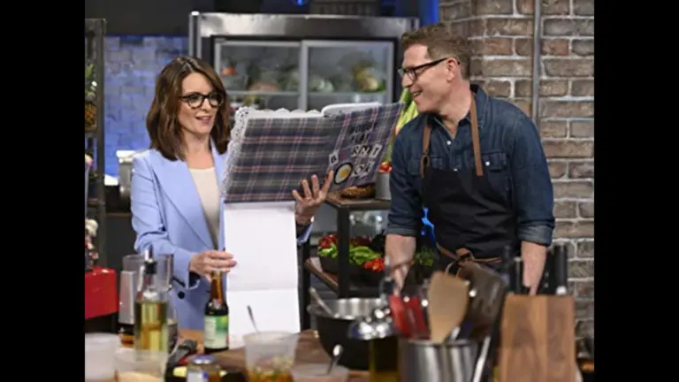 A woman in a blue blazer reading a large recipe book while a man in an apron looks on, both in a kitchen setting with various cooking ingredients and equipment around them. Tina Fey and Bobby Flay in Fey vs. Flay (2019)