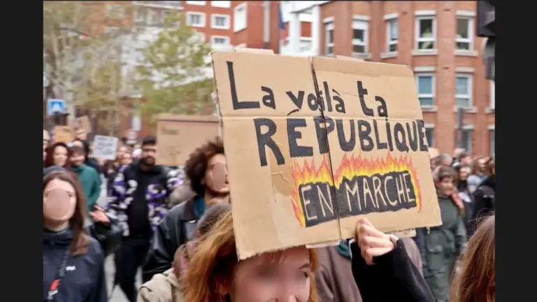A protester holding a sign that reads "La voilà ta RÉPUBLIQUE EN MARCHE" amidst a crowd of demonstrators in an urban setting.