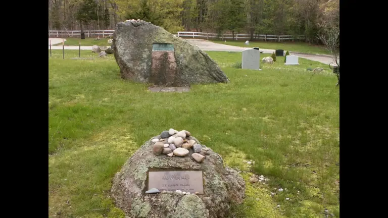 Jackson Pollock and Lee Krasner stones in Green River Cemetery in Springs, New York. Two large rocks in a grassy area, one with a plaque and a pile of small stones on top, the other appearing to have a carved surface. In the background, there are additional markers and trees.