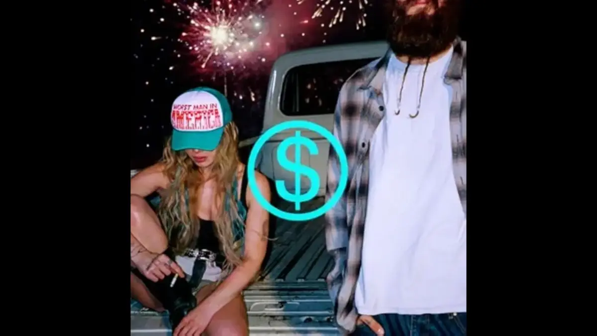Album cover for Slayyyter Wor$t Man in America - A woman sitting on a truck bed wearing a cap and a man standing next to her, with fireworks in the background and a dollar sign logo overlay.