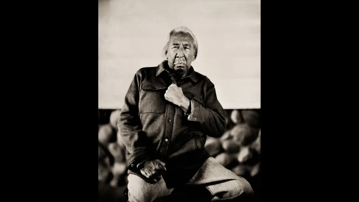 A black and white portrait of an elderly man with long gray hair and facial tattoos, sitting in front of a textured background, wearing a jacket and holding onto it with one hand. Leonard Peltier