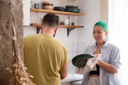A couple washes dishes together in a sunlit kitchen, sharing household chores and the mental load of running a home