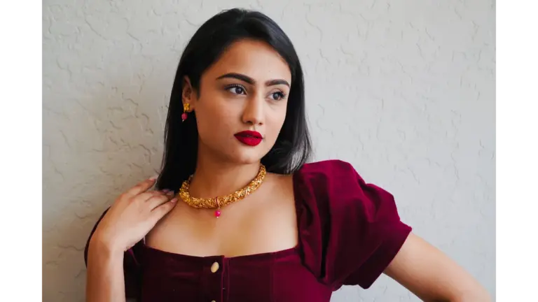 Stylish woman showcasing cherry cola makeup with rich red lipstick and gold jewelry, posing elegantly indoors for a glamorous beauty look.