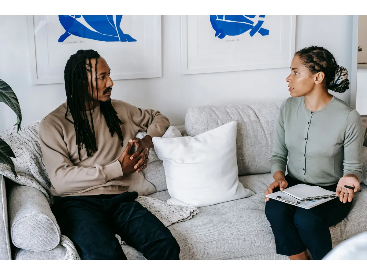 Two adults sit on a light-colored couch having a serious conversation, one holding a notebook and pen — a moment reflecting communication and adult kids independence.