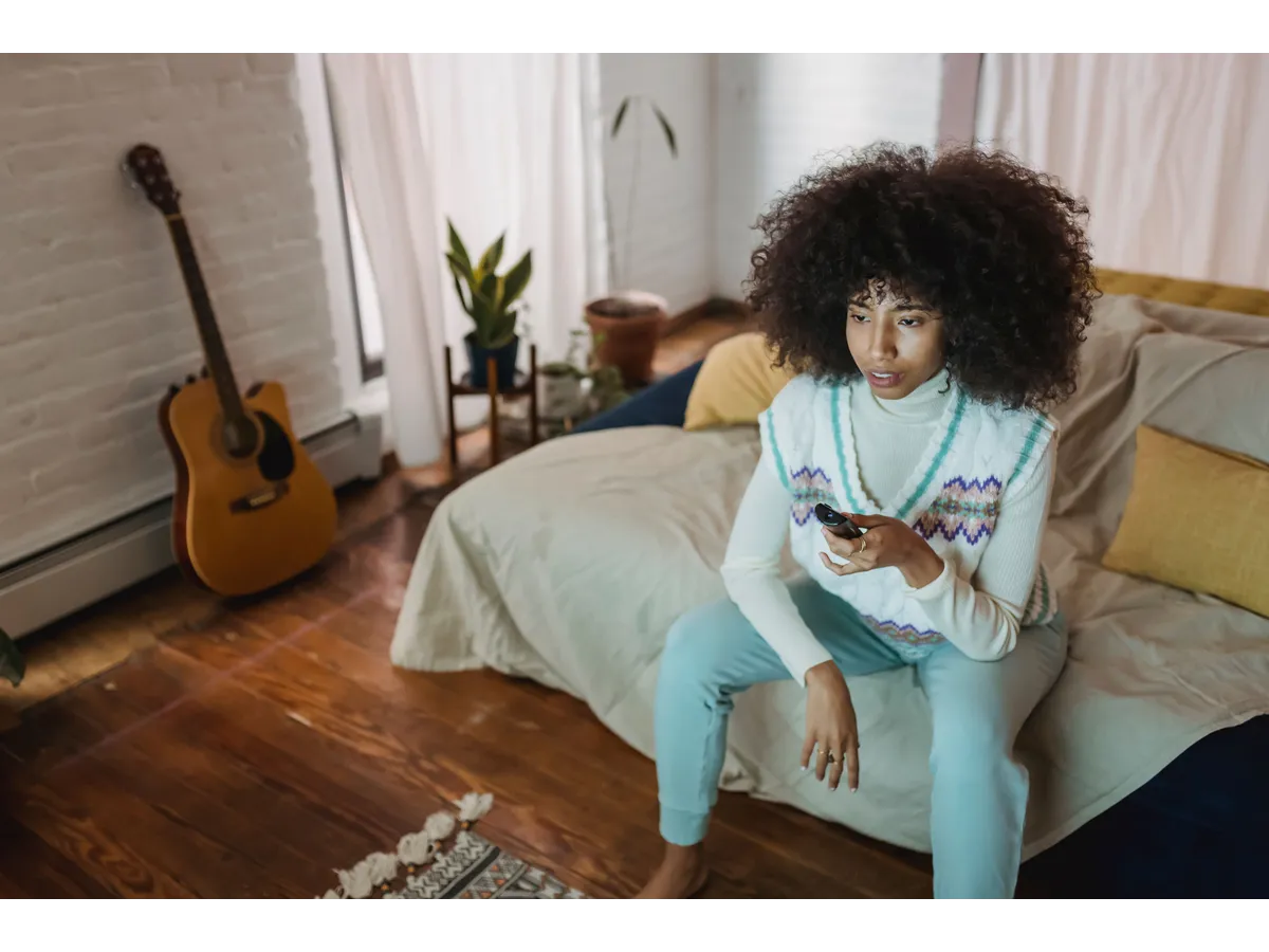 A woman with curly hair sits on a bed holding a remote, absorbed in a video moment that mirrors the introspective tone of Confessional Interviews.