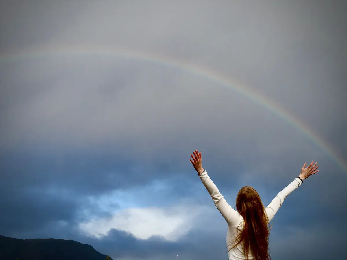 In today's daily devotional, a woman with outstretched arms looks at a sunrise and rainbow in the distance.
