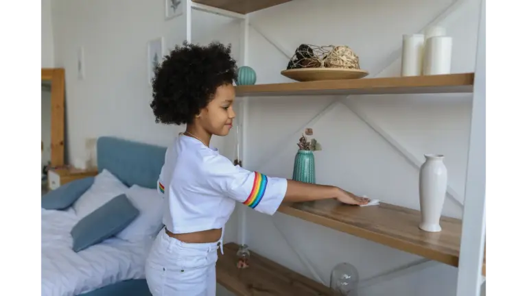 Young girl dusting a wooden shelf in a cozy, modern bedroom, showing everyday kids chore motivation in action.