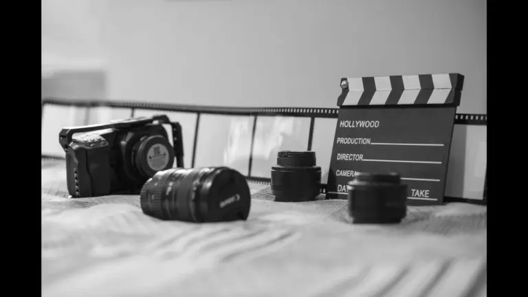 A black and white image featuring a camera, several camera lenses, and a film clapperboard on a bed with film strips in the background.